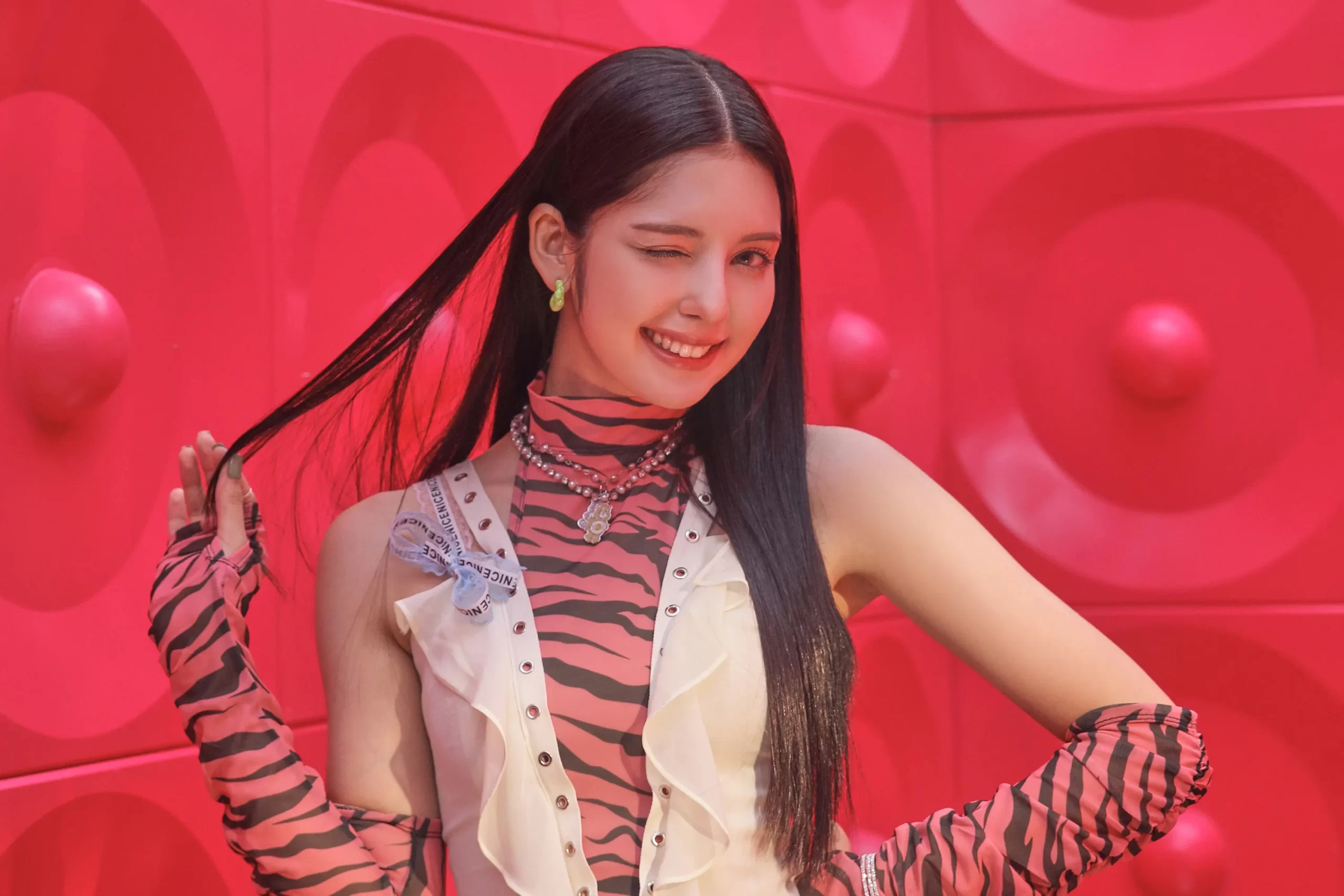 K-Pop girl Chanty Videla of Lapillus smiling and winking while holding a strand of her long black hair, wearing a pink and black zebra-print outfit with layered accessories, posing in front of a bright pink textured wall