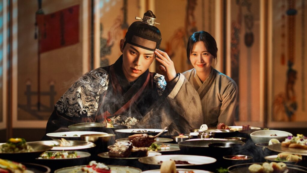 Korean actors Im Yoon-a and Lee Chae-min with food all around them in a promotional poster for ‘Bon Appétit, Your Majesty’, a K-drama for food lovers and rom-com fanatics
